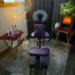 Massage chair positioned in a cozy room with a rug, fake fireplace, potted plant, folded towels, and tables holding oils, lotion bottles, and a lit decorative branch.