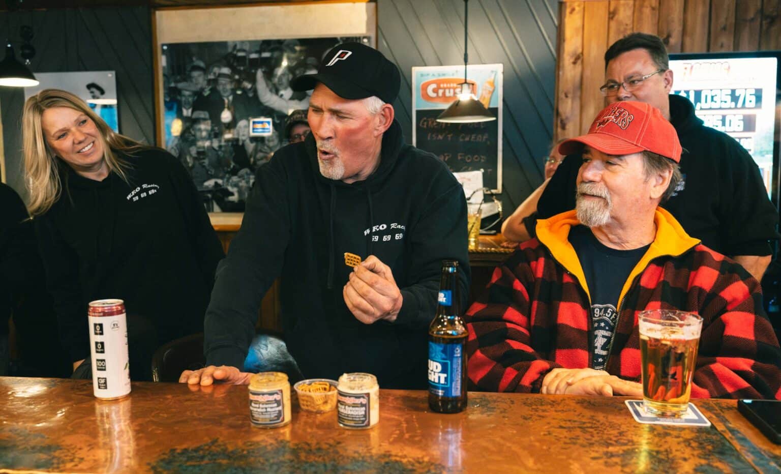 A group of people react as a man in a black hoodie eats something at a bar counter, with drinks and small food containers in front of them.