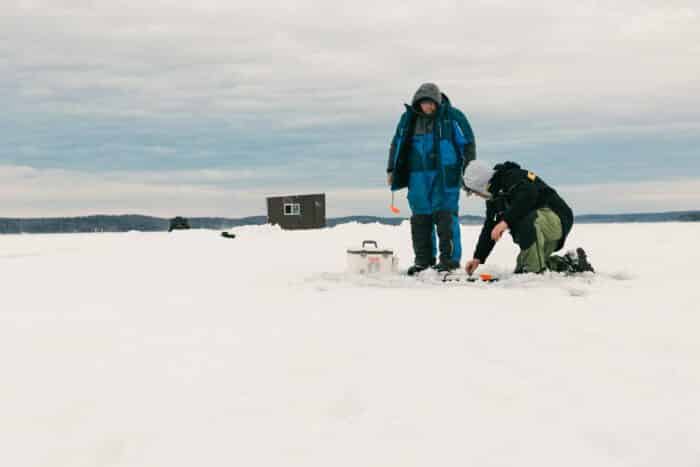 Two people are ice fishing on a snowy lake; one is kneeling by the fishing hole while the other stands nearby. An ice fishing hut and equipment are visible in the background.