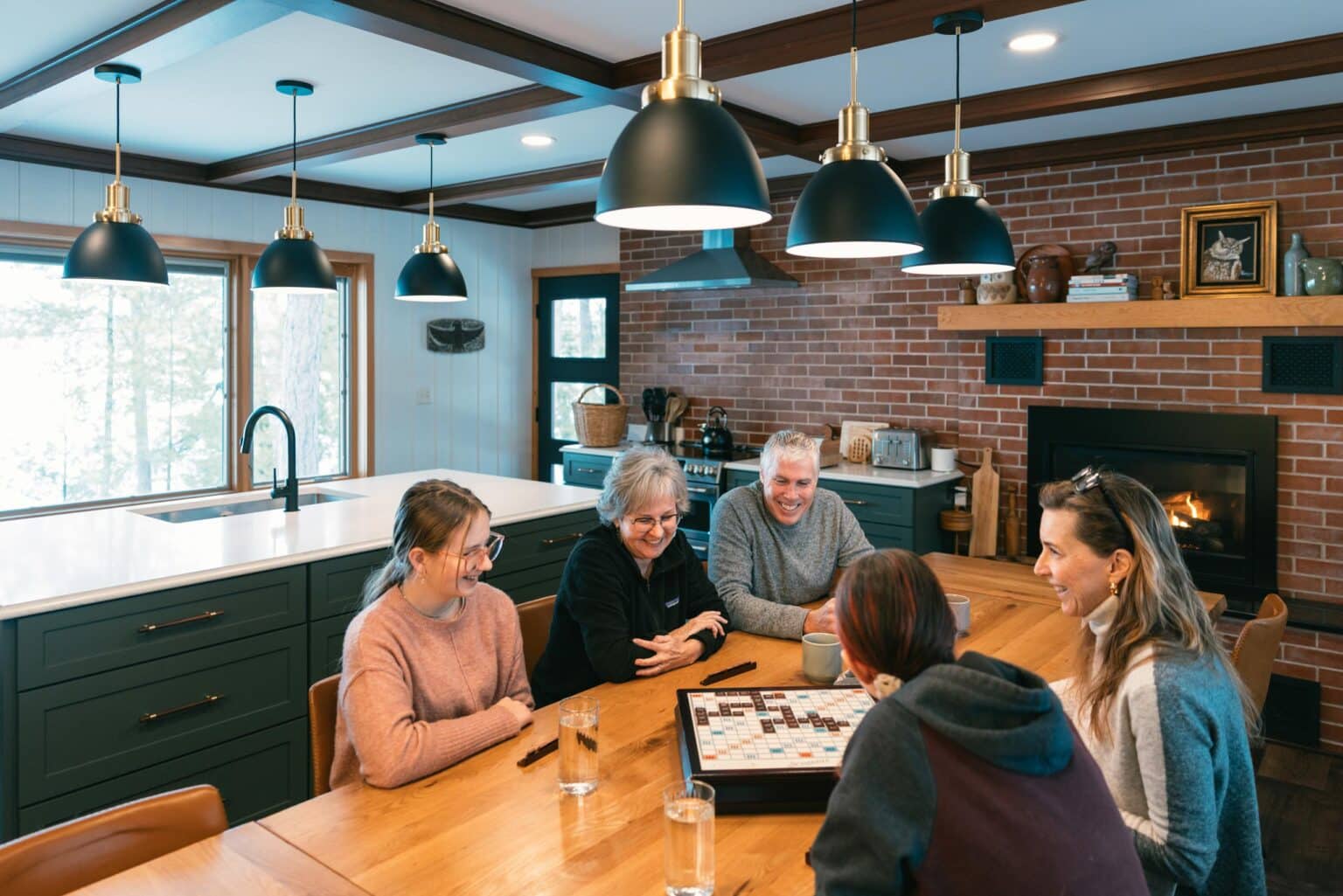 Five people sit around a wooden table in a kitchen, playing a board game and talking. Glasses of water are on the table and a fireplace is visible in the background.