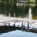 A metal and wood dock extends over a calm lake, with trees and grass visible on the opposite shore.