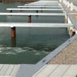 Empty boat docks with wooden piers extend over the water next to a gravel walkway lined with white posts.