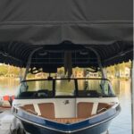A motorboat with brown and white seats is docked under a black canopy on a calm lake, with houses and trees visible in the background.