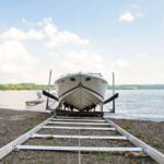 A white boat sits on a metal rail system at the edge of a lake, with a partly cloudy sky and distant shoreline in the background.
