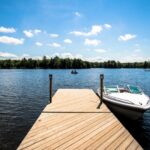 A white motorboat is docked at the end of a wooden pier extending into a calm lake, with trees lining the shoreline under a blue sky with scattered clouds.