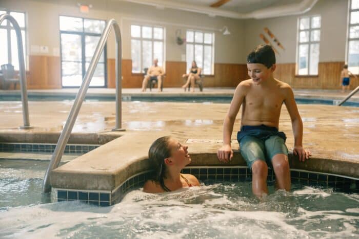 Two children talk and smile, one sitting on the edge and the other in a hot tub at an indoor pool facility. Adults sit in the background.