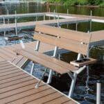 A wooden bench with armrests and cup holders is mounted on a dock overlooking a lake, with trees visible in the background.