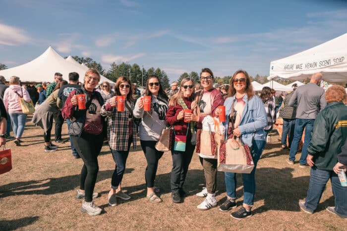 Six women stand outdoors at a festival, smiling and holding drinks, with tents and other people in the background under a clear sky.