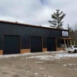 A black metal building with three large garage doors and a sign reading "Garage Heroes," with a white SUV parked outside on a snowy ground.