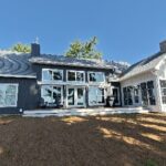 A modern two-story house with large windows, dark exterior siding, and a stone chimney, surrounded by trees under a clear blue sky.