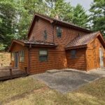 A two-story wooden cabin with brown siding is surrounded by trees, featuring several windows and a small deck in the front.