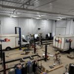 Two FedEx Ground delivery trucks are parked inside a large, well-lit garage or workshop with tools, equipment, and automotive parts scattered around.