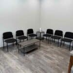 A small waiting room with seven black chairs along the wall, a rectangular coffee table in the center, and a small table with a tissue box in the corner.