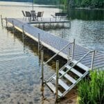 A lakeside dock with metal steps leads to a patio area with a table, chairs, and a blue umbrella; surrounded by calm water and trees.
