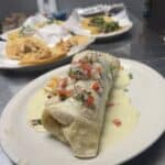 A burrito topped with cheese sauce, diced tomatoes, and herbs on a white plate, with a plate of tacos and sides blurred in the background.