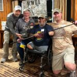 Four men pose indoors in front of a VWWA banner; one is seated in a wheelchair holding a fishing rod, the others stand, smiling, with fishing gear and event badges visible.