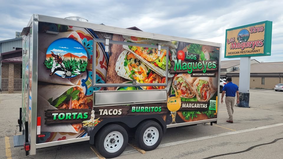 Food truck for Los Magueyes Mexican Restaurant featuring images of tortas, tacos, and burritos, parked in a lot with a man walking nearby and the restaurant sign visible.
