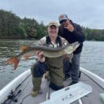 Two men on a boat; one kneels and holds a large fish, while the other stands behind making a hand gesture. Trees and water are visible in the background.