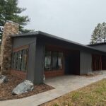 A modern, single-story house with dark siding, large windows, a stone chimney, and a paved walkway surrounded by trees and grass.