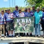 A group of people poses outdoors by a lake, many wearing matching shirts, holding a large banner with a deer skull and antlers logo.