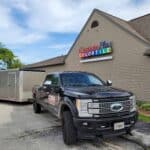 A black Ford pickup truck with a trailer is parked outside a CarpetsPlus COLORTILE store on a cloudy day.