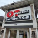 Exterior sign for "The Overtime Sports Bar and Grill" mounted above the entrance door of a beige building with the address number 175.