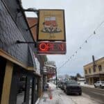 A sidewalk view of Brothermanz Bar with a lit "OPEN" sign on a snowy street lined with parked cars and buildings.