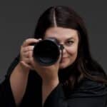 A person with long dark hair points a camera toward the viewer, partially obscuring their face, against a plain dark background.