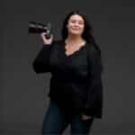 A woman in a black blouse and jeans poses holding a camera over her shoulder, standing against a plain dark background.