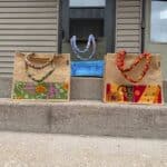 Three reusable fabric bags with colorful patterned handles and fronts are placed on concrete steps outside a building with siding and a closed door.