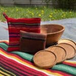 A wooden bowl, four wooden coasters, a brown leather pouch, and a red woven pot holder are arranged on a colorful striped blanket outdoors.