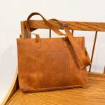 A brown leather tote bag with dual handles and a shoulder strap rests on a wooden bench.