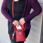 A person wearing a black dress and purple cardigan places a white smartphone into a red crossbody bag while standing outside near a wall.