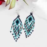 A pair of beaded dangle earrings with a geometric pattern in blue, turquoise, black, and white, displayed on a white surface with a blurred floral background.