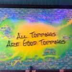 A colorful mural on a brick wall displays the phrase "All Toppings Are Good Toppings" in bold black lettering above a row of booth seating.