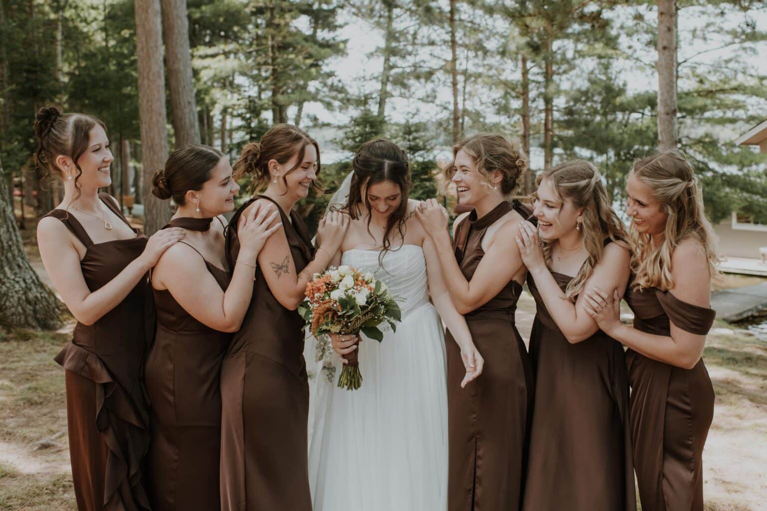 A bride in a white wedding dress stands surrounded by six bridesmaids in brown dresses outdoors in a wooded area.