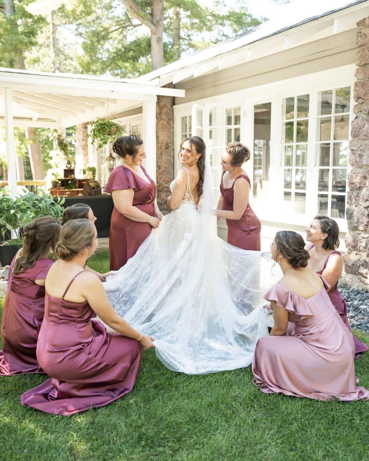 A bride in a white wedding gown smiles over her shoulder while her bridesmaids in mauve dresses arrange her long lace veil on the grass.
