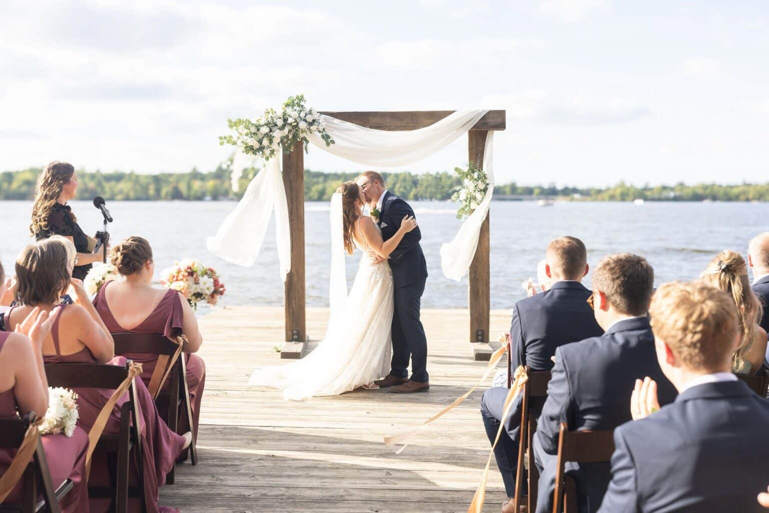 A bride and groom kiss under a floral arch on a wooden dock during their wedding ceremony, with guests seated on either side.
