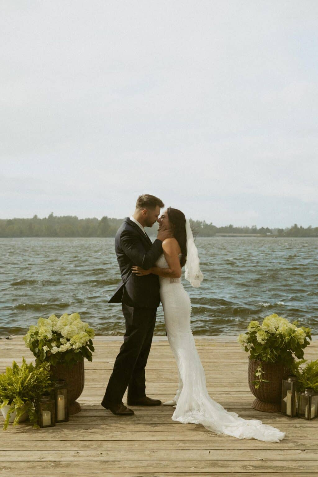 A bride and groom face each other on a wooden dock by a lake.