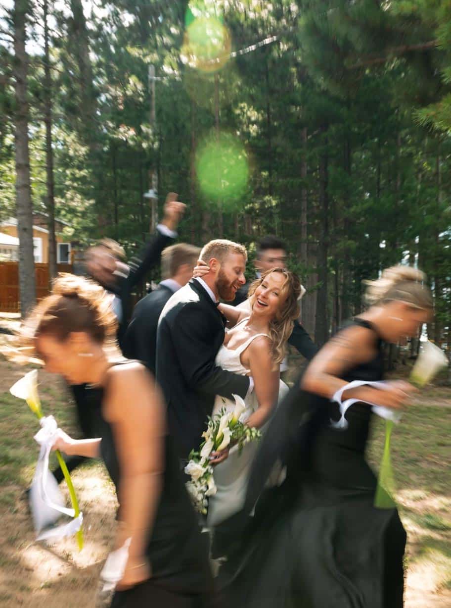 A bride and groom embracing in the center of a group of wedding guests, captured in motion in a wooded area.