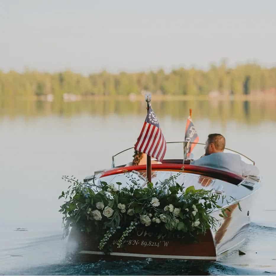 A couple sitting in a wooden boat decorated with white flowers on a calm lake, with an American flag attached to the stern.