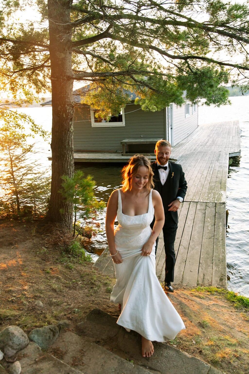 A bride in a white gown and a groom in a black tuxedo walking from a wooden dock toward the shore.