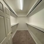 Empty walk-in closet with beige carpet, white walls, two long shelves and hanging rods on both sides, and a fluorescent ceiling light.