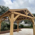 A wooden pergola with a peaked roof stands on a stone patio in a landscaped backyard, with houses and trees in the background under a cloudy sky.