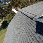 A gray shingled roof with a ridge vent and ventilation pipe, surrounded by trees and grass, viewed from above.