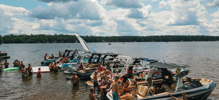 Several boats are docked side by side on a lake, with groups of people in swimsuits sitting on the boats and swimming near the shore under a partly cloudy sky.
