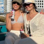 Two women sitting close together on a boat, both wearing sunglasses, hats, light-colored outfits, and holding canned drinks, smiling at the camera.