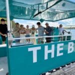 A group of people stand on a teal boat named "THE BUB," docked by a lake with trees and blue sky in the background.