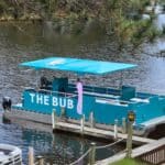 A turquoise boat with a canopy labeled "THE BUB" is docked at a wooden pier on a calm lake, surrounded by trees and other small boats.