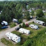 Aerial view of an RV park with several trailers and vehicles parked among trees, surrounded by greenery on a sunny day.